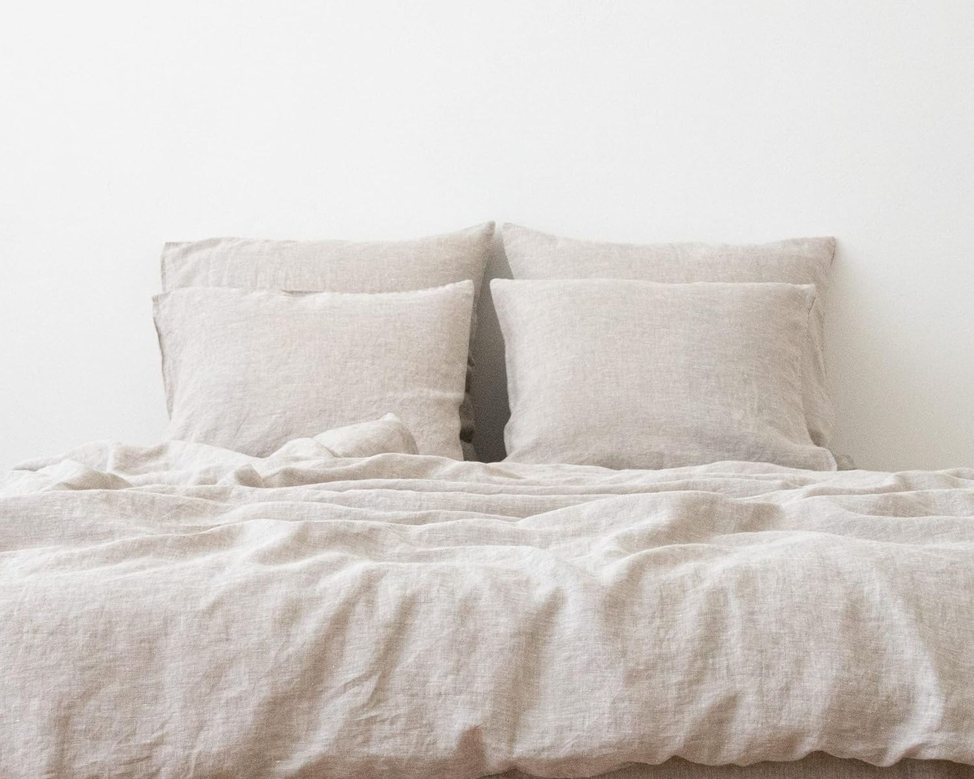 A bed made up in natural washed linen, with four linen pillows against a white wall